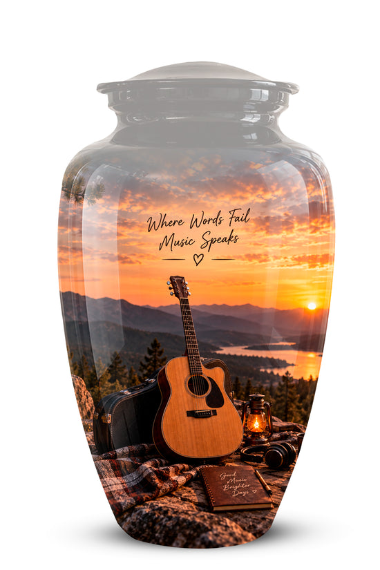 A clear glass urn with a lid, featuring a printed scenic design of an acoustic guitar, a lantern, and a book on a blanket in front of a mountain sunset over water. The text 'Where Words Fail Music Speaks' is printed on the front.