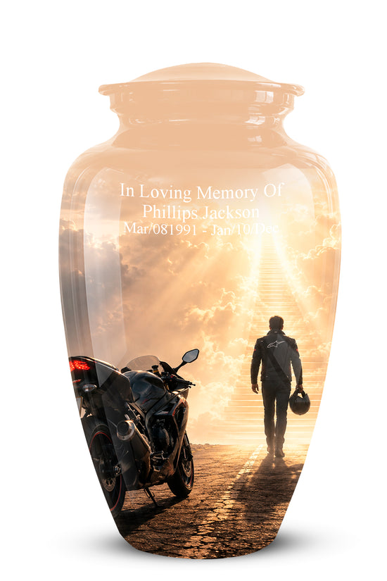 A clear glass memorial urn with a lid, featuring a printed sunset scene of a motorcycle and a person walking toward a bright horizon. White engraved text on the front reads, 'In Loving Memory Of Phillips Jackson' with dates below, shown on a white background.