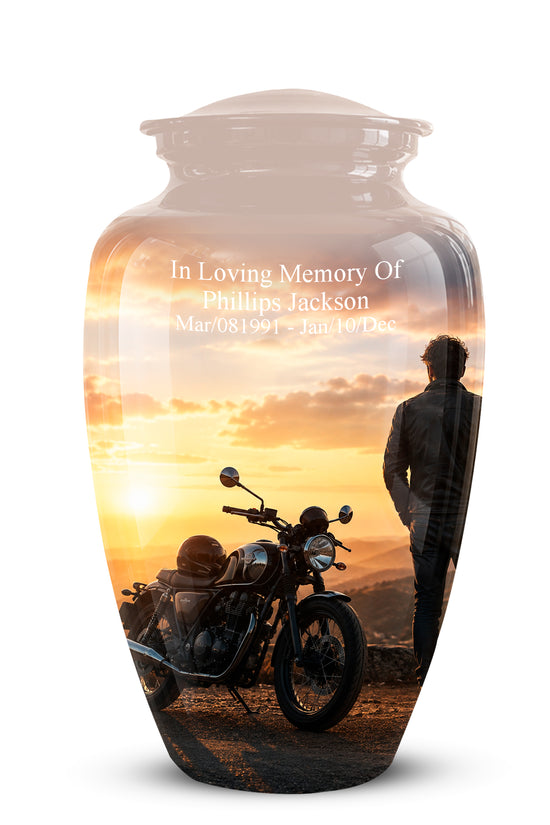 A personalized glass urn with a lid, featuring a printed sunset landscape design of a man and a motorcycle. The front of the urn has white text reading 'In Loving Memory Of Phillips Jackson' with dates below, shown on a clean white background.
