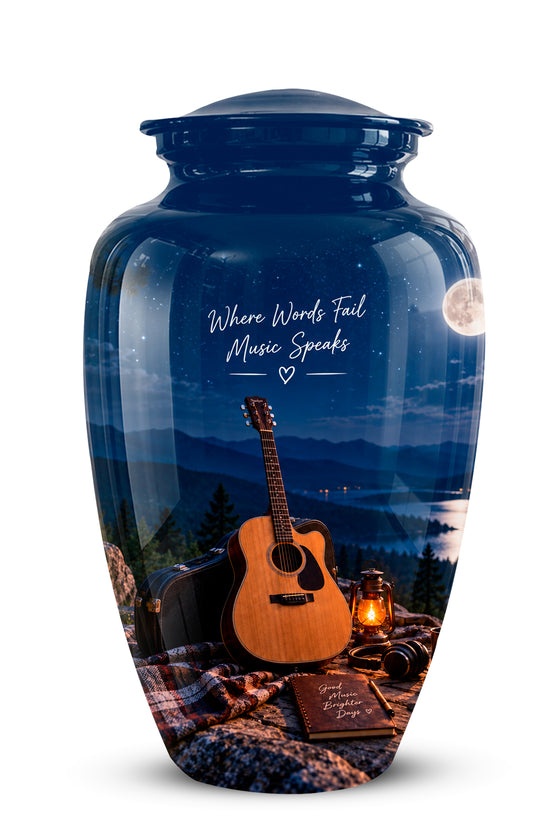 A glossy blue ceramic cremation urn featuring a scenic landscape print of a mountain lake at night with an acoustic guitar, lantern, and blanket. The urn has a matching blue lid and white script text that reads 'Where Wounds Heal Music Speaks'.