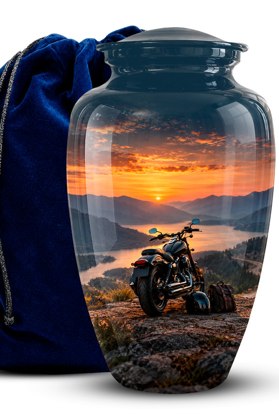 A glossy glass urn with a scenic sunset landscape print showing mountains, a lake, and a motorcycle, displayed next to a dark blue velvet drawstring bag on a white background.