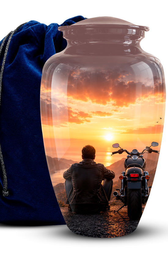 A clear glass cremation urn with a lid, printed with a sunset scene showing a person sitting next to a motorcycle, displayed next to a dark blue velvet drawstring bag on a white background.