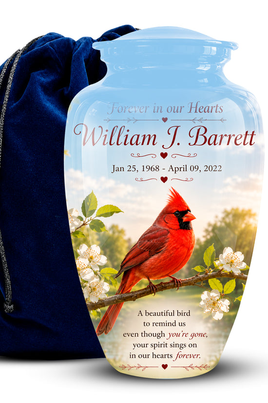 Personalized ceramic memorial urn with a blue neck and a printed photo of a red cardinal on a branch, shown next to a dark blue drawstring bag. The urn features the text 'Forever in our Hearts William J.
