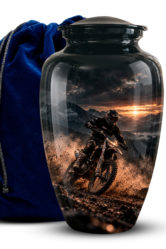 A glossy black ceramic cremation urn with a custom printed graphic of a dirt bike rider on a mountain road at sunset, shown next to a royal blue velvet drawstring bag on a white background.
