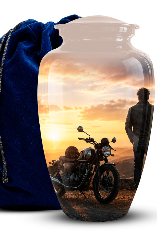 A clear glass urn-shaped vase with a printed scenic photo of a motorcycle and a person at sunset, shown next to a blue velvet drawstring bag on a white background.
