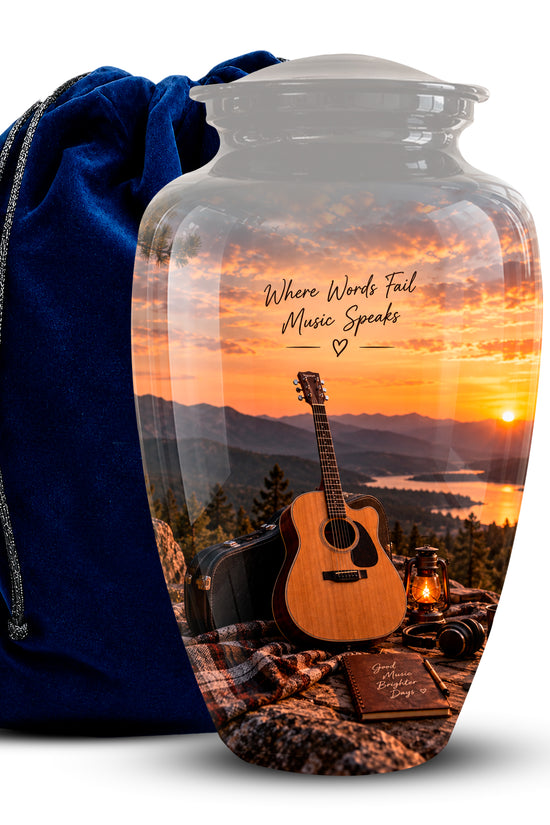A clear glass urn-shaped vase with a printed scenic sunset design showing an acoustic guitar, mountains, lake, and the phrase 'Where Words Fail Music Speaks,' displayed next to a dark blue velvet drawstring bag on a white background.