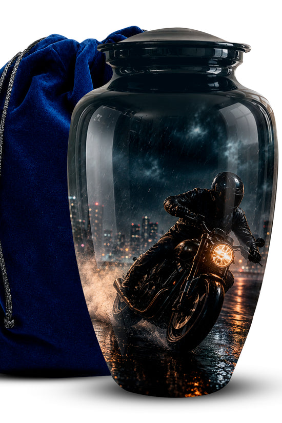 A glossy black cremation urn with a lid, featuring a printed graphic of a motorcyclist riding at night, shown next to a blue velvet drawstring bag on a white background.