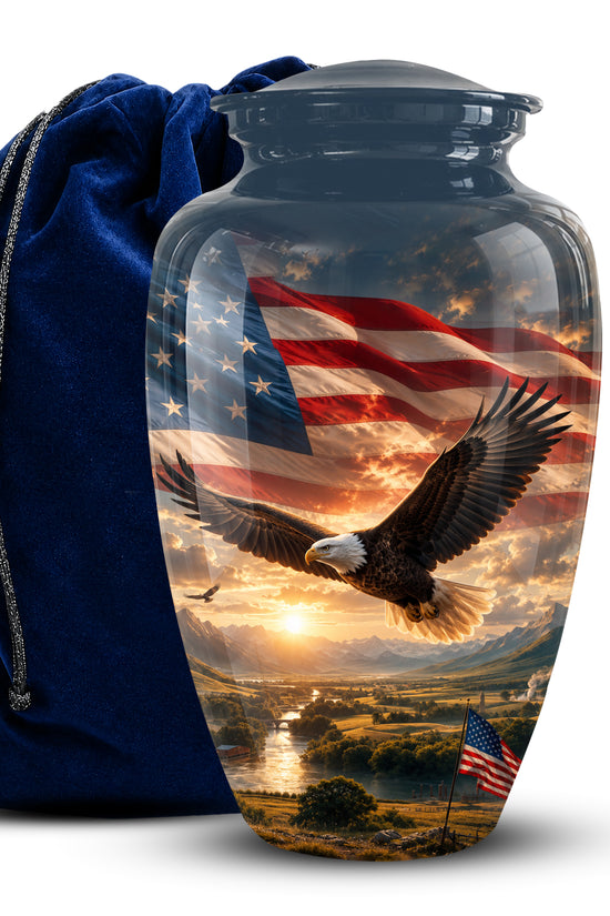 A clear glass cremation urn with a dark gray lid, featuring a printed design of a bald eagle flying over a landscape beneath an American flag. A dark blue velvet drawstring bag is positioned behind the urn on a white background.