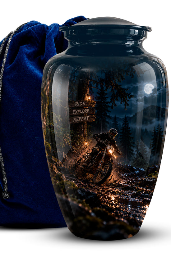 A glossy black ceramic cremation urn with a lid, featuring a printed scenic graphic of a mountain biker at night on a forest trail. A dark blue velvet drawstring bag is positioned behind the urn on a white background.