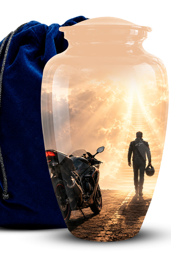 A clear glass cremation urn with a printed scenic design of a motorcycle and a person walking toward a bright sunset, shown next to a dark blue velvet drawstring bag on a white background.