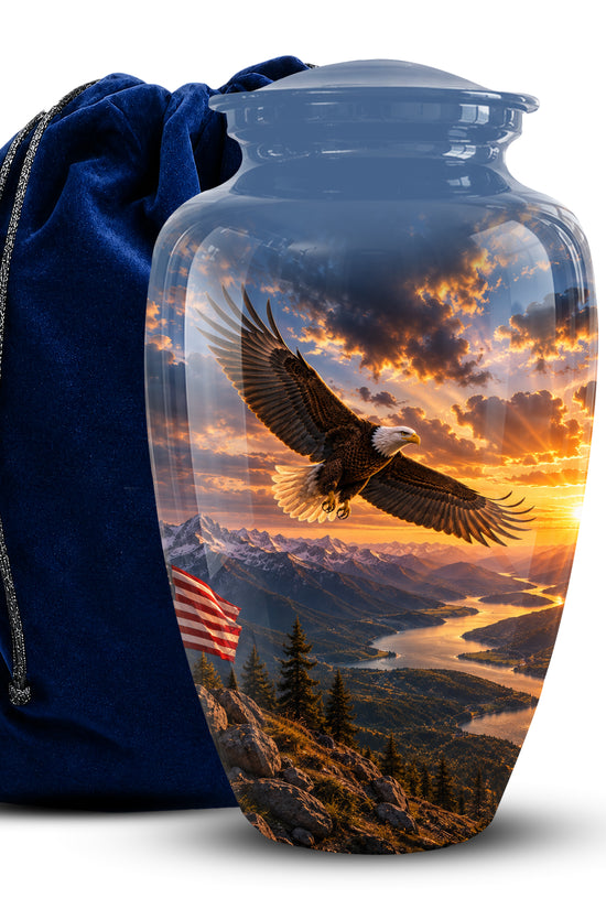 A glossy ceramic cremation urn with a lid, featuring a printed scenic design of a bald eagle flying over mountains and a river at sunset, shown next to a dark blue velvet drawstring bag on a white background.