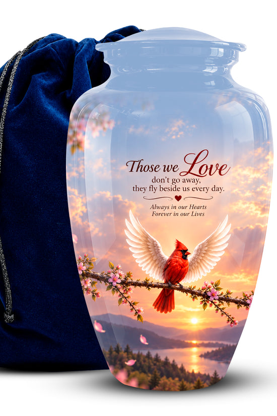 A clear glass cremation urn vase with a printed sunset scene featuring a red cardinal on a branch, placed in front of a dark blue velvet drawstring bag. The vase has the printed text 'Those we Love don't go away, they fly beside us every day.
