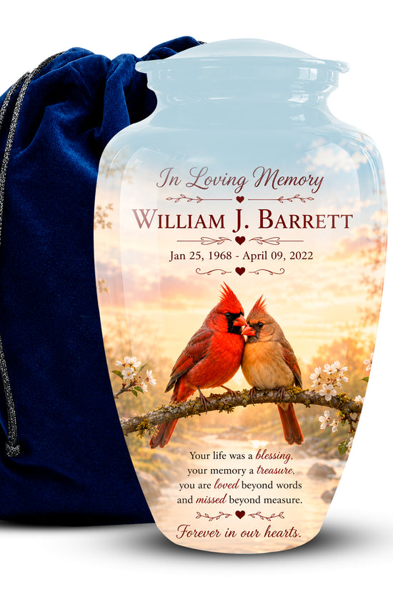 Personalized clear glass urn with a printed memorial design featuring two cardinals on a branch at sunset, shown next to a dark blue velvet drawstring bag on a white background.