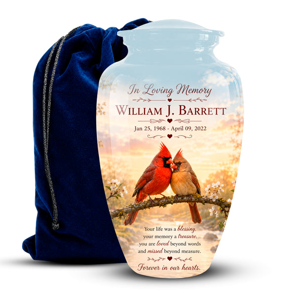 Personalized clear glass urn with a printed memorial design featuring two cardinals on a branch at sunset, shown next to a dark blue velvet drawstring bag on a white background.