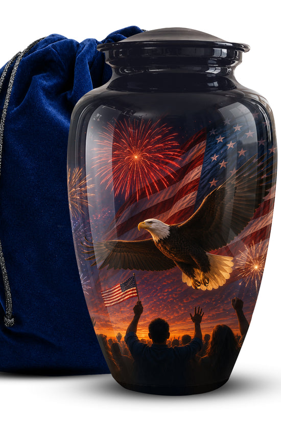 A glossy black cremation urn with a lid, featuring a patriotic print of an eagle flying in front of an American flag and fireworks, shown next to a royal blue velvet drawstring bag on a white background.