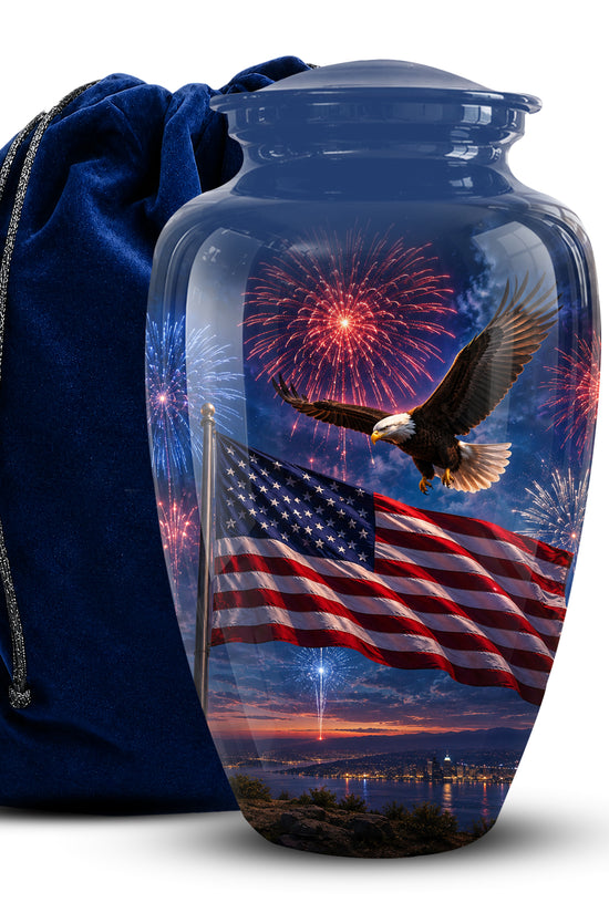 A clear glass cremation urn with a lid, printed with an American flag, bald eagle, fireworks, and sunset landscape. A dark blue velvet drawstring bag stands behind it on a white background.