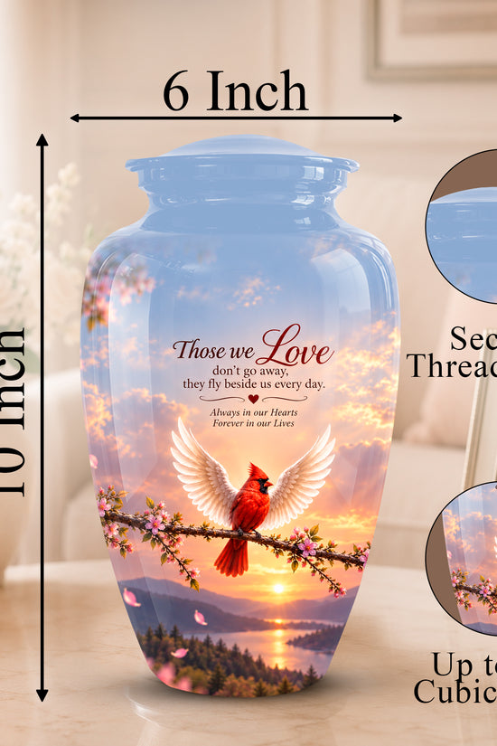 A printed ceramic cremation urn shown on a tabletop with dimension arrows indicating 10-inch height and 6-inch width. The urn has a blue threaded lid and a scenic print of a red cardinal on a branch at sunset, with memorial text reading 'Those we Love.