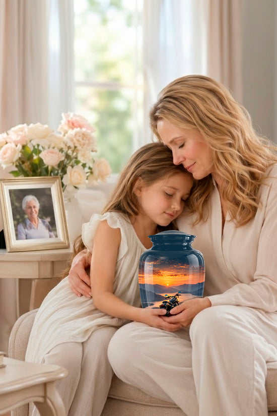 A woman and a young girl sit together on a light beige sofa in a bright living room, holding a small blue glass jar that contains a colorful sunset landscape. A framed photo of an elderly woman and a vase of pink flowers sit on a side table nearby.