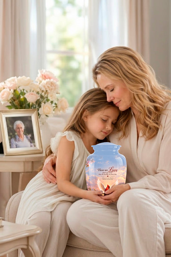 A woman and a young girl sit together on a beige sofa in a bright living room, holding a glowing personalized urn. The urn is light blue with a sunset illustration and the words 'You Are Loved' and 'Love, Mom'.