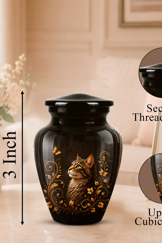 Black cremation urn with a glossy finish, featuring a detailed illustration of a tabby cat and gold floral scrollwork. The urn is shown on a marble tabletop in a warm, softly lit living room setting, with dimension and feature callout overlays.