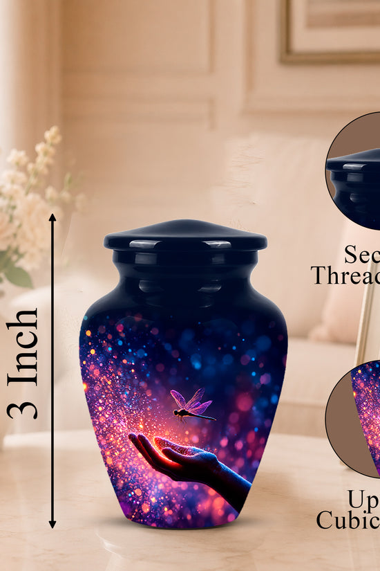 A small urn-shaped vase with a dark blue lid and a colorful cosmic print featuring a glowing hand, dragonfly, and purple-blue bokeh lights. The image includes a height marker reading 3 Inch and two circular inset callouts showing the secure threaded lid and up to 3 cubic inches capacity, set in a softly lit indoor room.