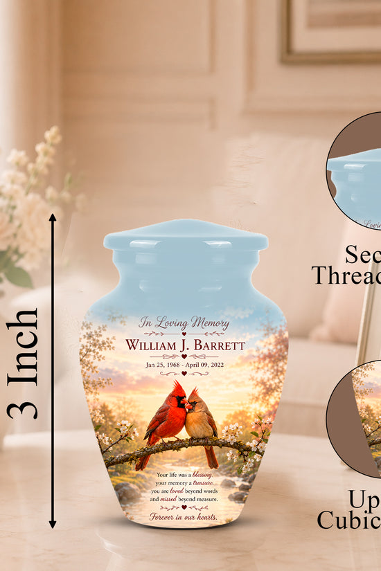 Personalized ceramic memorial urn with a light blue threaded lid, featuring a printed design of two cardinals on a branch with cherry blossoms and memorial text. The urn is shown on a tabletop with a blurred home interior background, including a vase of flowers and framed photo.