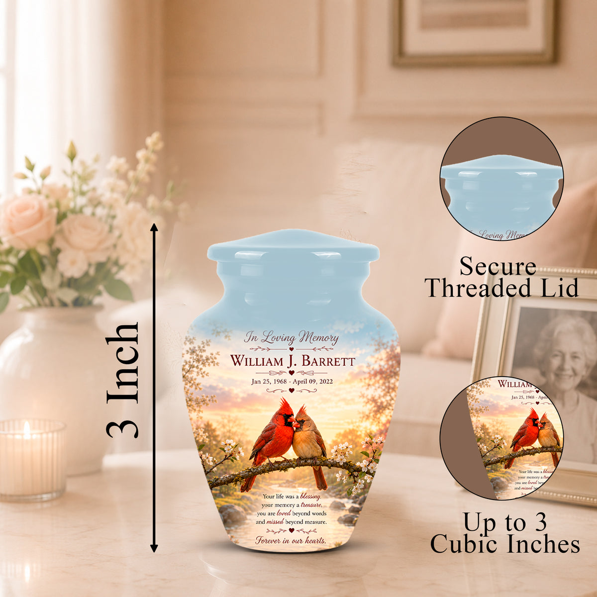 Personalized ceramic memorial urn with a light blue threaded lid, featuring a printed design of two cardinals on a branch with cherry blossoms and memorial text. The urn is shown on a tabletop with a blurred home interior background, including a vase of flowers and framed photo.