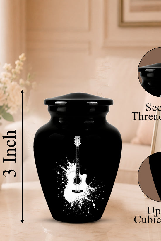 Black ceramic urn with a white guitar graphic and size measurement overlay, shown on a tabletop with decorative flowers and framed photo in a softly lit room. Callout circles highlight the secure threaded lid and up to 3 cubic inch capacity.
