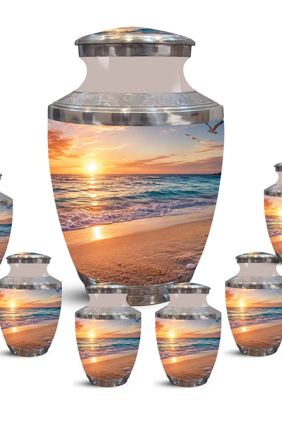 A set of cremation urns displayed on a plain white background, including one large urn and five smaller matching urns. The urns have a glossy finish with a printed beach sunset scene and silver metallic bands near the top.
