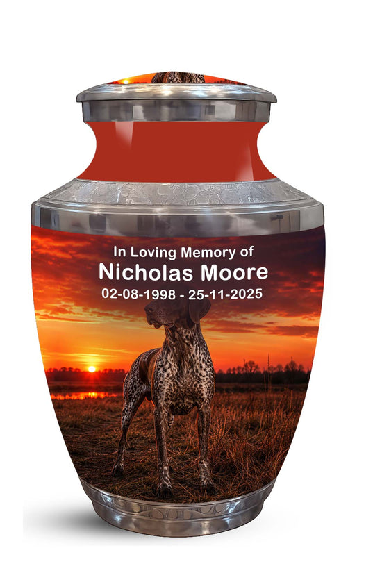 A memorial urn with a glossy red neck and silver metallic top and base, featuring a printed photo of a greyhound dog at sunset with personalized text reading 'In Loving Memory of Nicholas Moore' and dates.