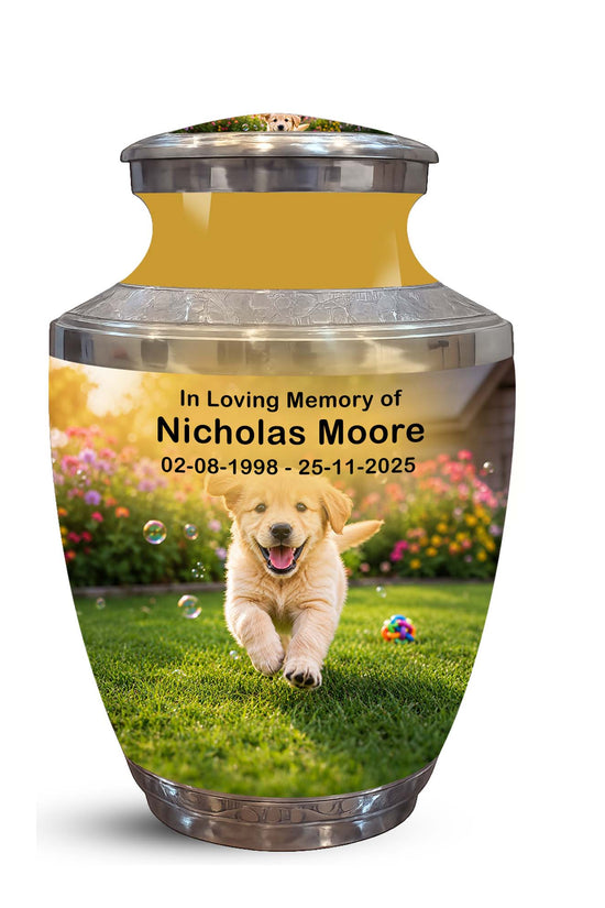 A memorial urn with a printed photo of a golden retriever puppy on a grassy lawn, with the text 'In Loving Memory of Nicholas Moore 02-08-1998 - 25-11-2025'. The urn has a metallic gold interior, a silver metallic lid and base, and is shown on a plain white background.