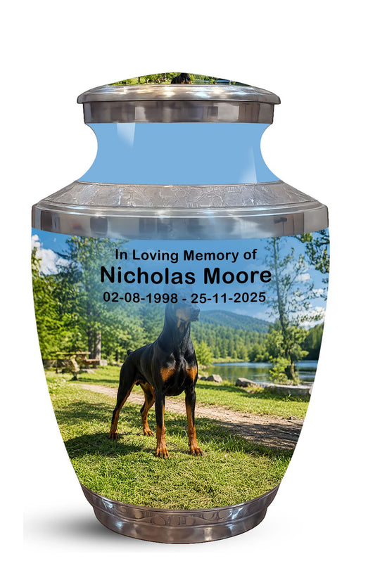 A memorial urn with a custom printed photo of a black and tan dog on a grassy lawn by a lake, with the text 'In Loving Memory of Nicholas Moore 02-08-1998 - 25-11-2025'. The urn has a metallic silver top and bottom and a light blue upper section.