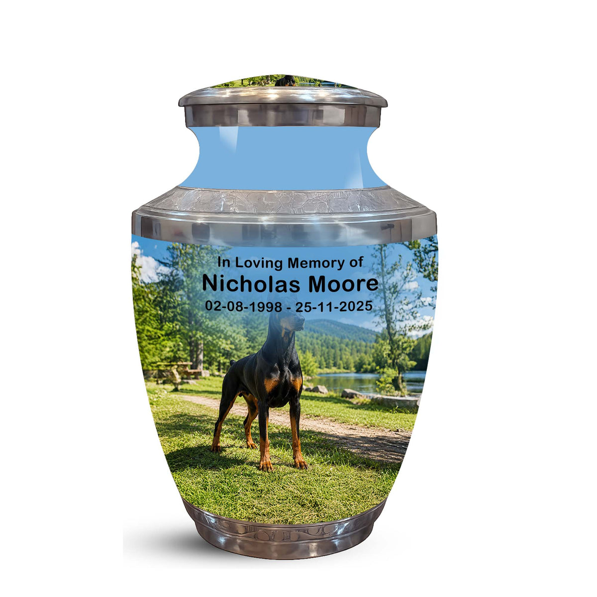 A memorial urn with a custom printed photo of a black and tan dog on a grassy lawn by a lake, with the text 'In Loving Memory of Nicholas Moore 02-08-1998 - 25-11-2025'. The urn has a metallic silver top and bottom and a light blue upper section.