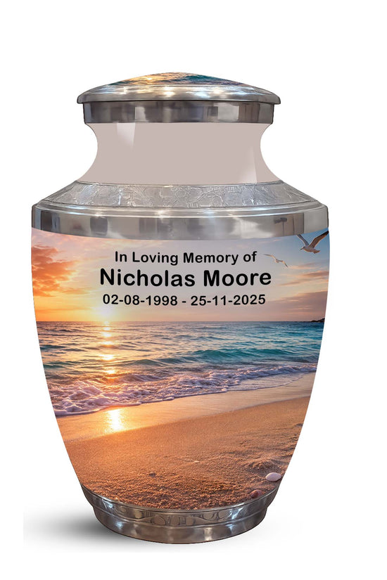 A memorial urn with a lid, featuring a printed beach sunset photo on the front and personalized text reading 'In Loving Memory of Nicholas Moore 02-08-1998 - 25-11-2025'. The urn has a silver metallic base and rim with a frosted upper section.