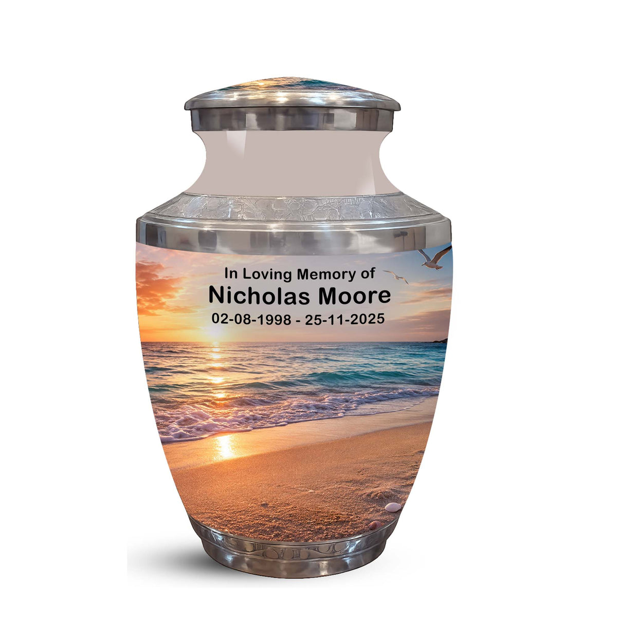 A memorial urn with a lid, featuring a printed beach sunset photo on the front and personalized text reading 'In Loving Memory of Nicholas Moore 02-08-1998 - 25-11-2025'. The urn has a silver metallic base and rim with a frosted upper section.