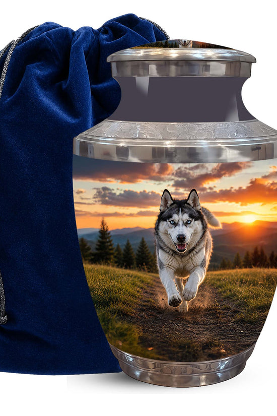 A stainless steel cremation urn with a custom printed photo of a husky running across a field at sunset, shown next to a dark blue velvet drawstring bag on a white background.
