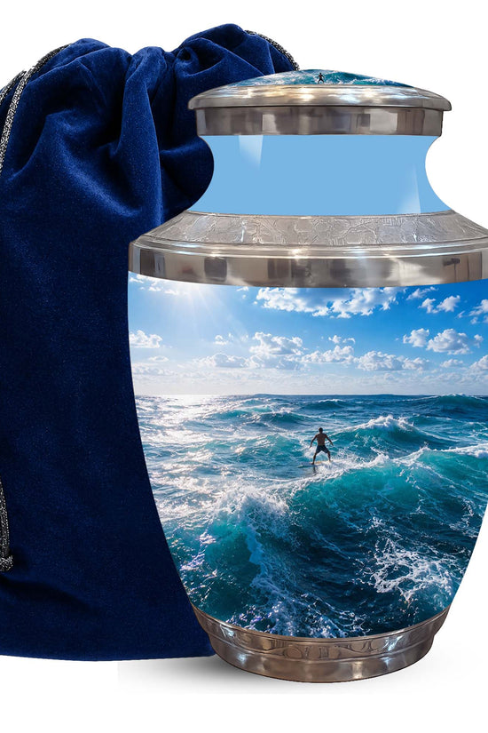 A ceramic cremation urn with a custom printed ocean wave scene featuring a surfer, shown next to a dark blue velvet drawstring bag on a white background.