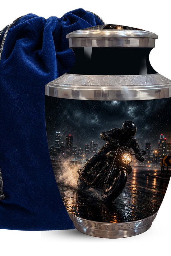 A metallic urn-shaped keepsake or cremation container with a printed motorcycle rider at night design, shown next to a dark blue velvet drawstring bag on a white background.