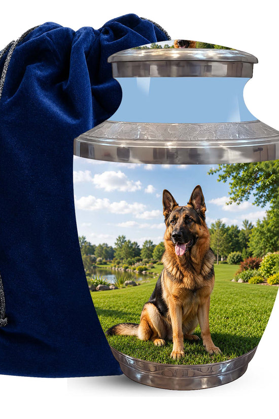 A pet memorial urn shaped like a glass jar with a silver metal lid and base, printed with a photo of a German Shepherd sitting on grass under a blue sky. A dark blue velvet drawstring bag is placed beside it on a white background.