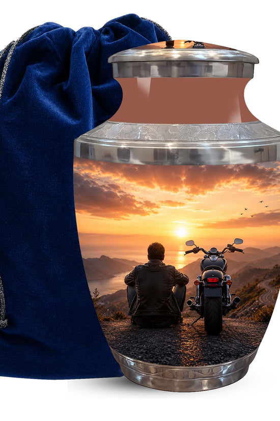 A metallic cremation urn with a lid, featuring a printed scenic image of a person sitting on a motorcycle at sunset, next to a dark blue velvet drawstring bag on a white background.