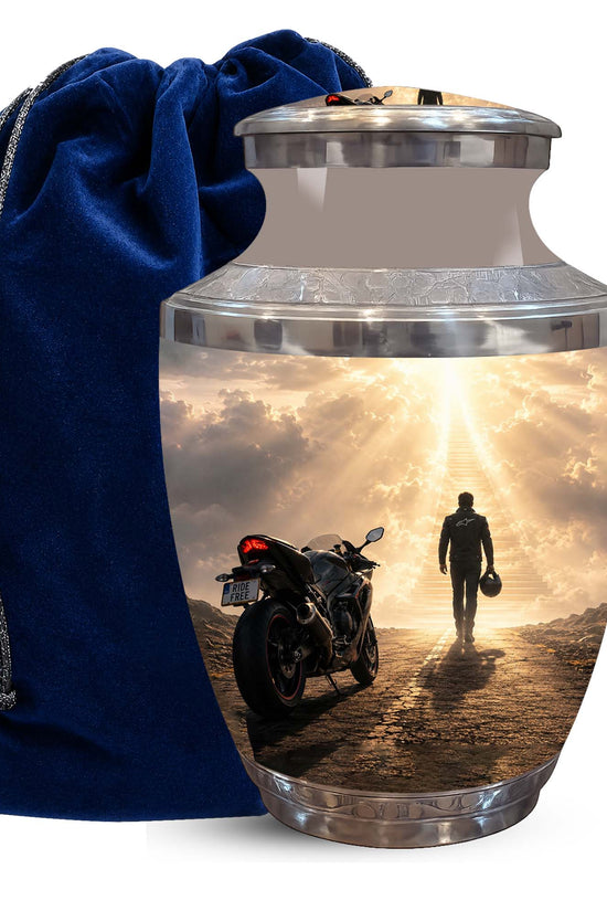 A metallic cremation urn with a custom printed photo of a motorcycle and a person walking toward a bright sky, shown next to a dark blue velvet drawstring bag on a white background.