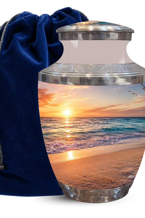 A ceramic cremation urn with a silver-tone lid and base, featuring a printed beach sunset design, shown next to a dark blue velvet drawstring bag on a white background.