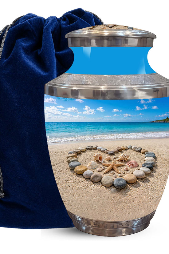 A ceramic urn with a stainless steel lid and base, featuring a printed beach scene with a heart-shaped arrangement of seashells and starfish on the sand. A dark blue velvet drawstring bag is positioned behind the urn on a white background.
