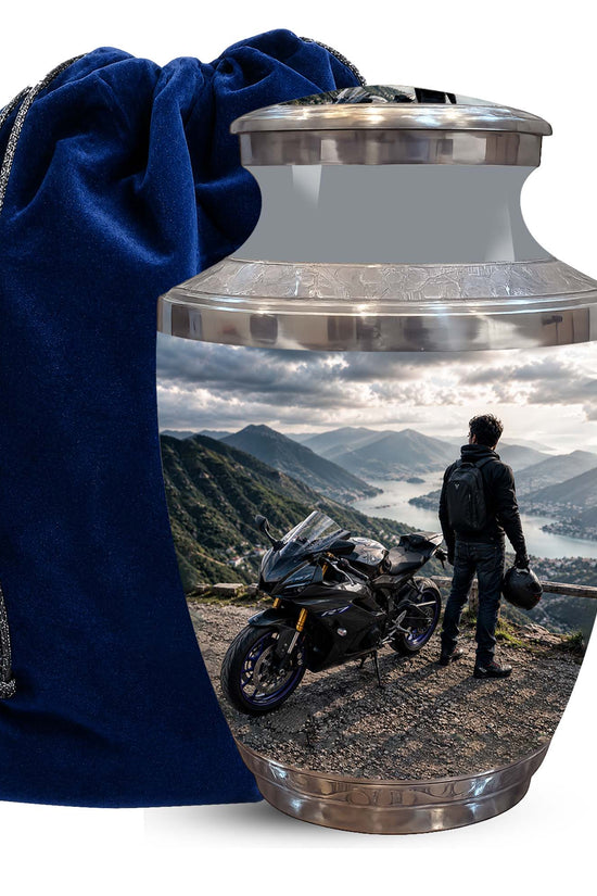 A glossy metallic cremation urn with a custom landscape photo print of a motorcyclist on a mountain road, shown next to a dark blue velvet drawstring bag on a white background.