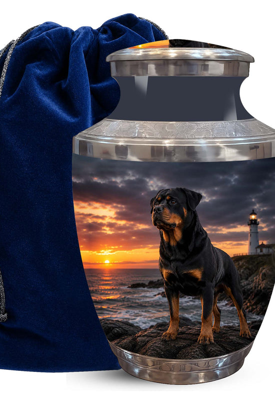 A memorial urn with a printed photo of a Rottweiler dog at sunset by the ocean, shown next to a dark blue velvet drawstring bag on a white background.